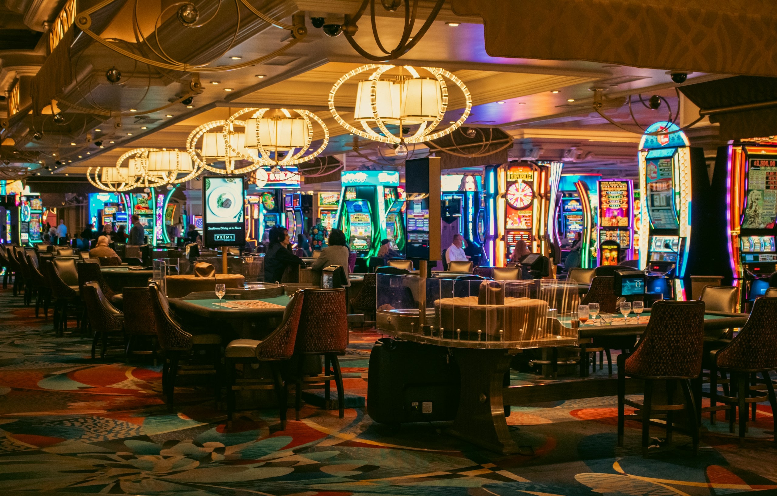 A casino floor of gaming tables and machines stretching away beneath chandeliers.