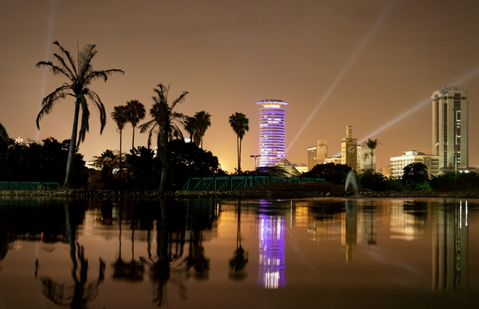 The Nairobi skyline at dusk, reflected in water beyond a line of trees.